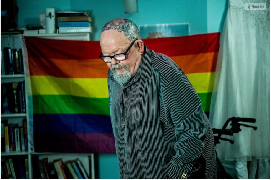 Image of Geoff in front of a rinabnow flag. Geoff has a tatood head, glasses and goatee. He is wearing a grey button up shirt